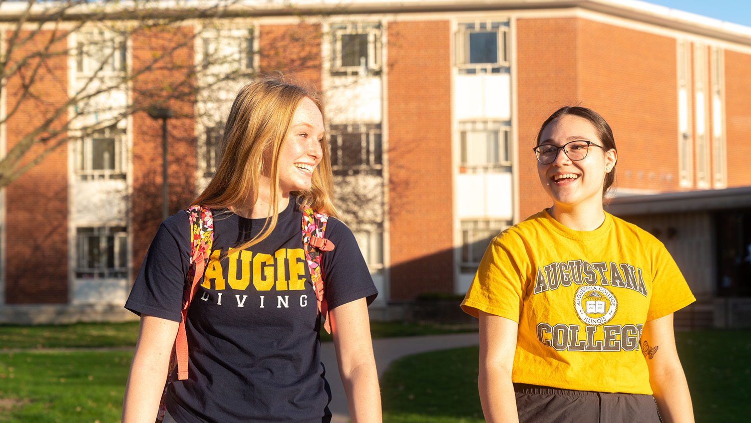 Two students wearing Augustana shirts walk and talk between residence halls