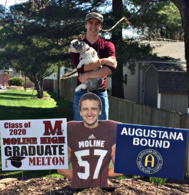 John standing behind 3 signs. One of the signs is a photo of him in a maroon football jersey