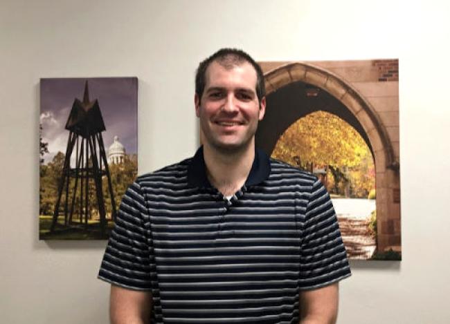Ryan (Man) standing tall in front of 2 pictures. Wearing a blue shirt with horizontal stripes.