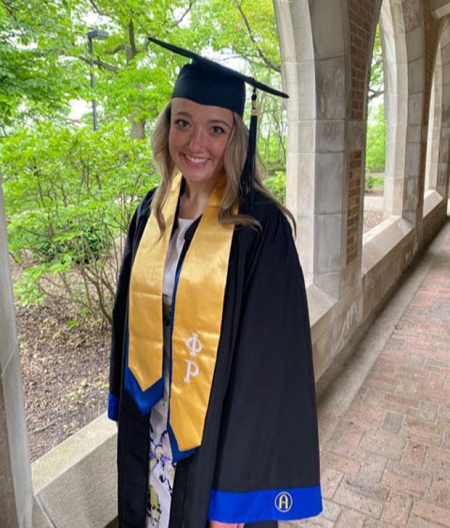 blonde woman in cap and gown. Tree and leaves in the background