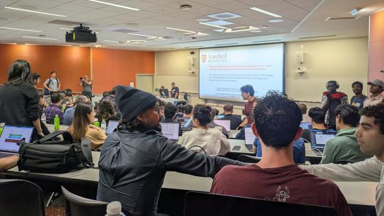 a wide shot of a classroom at Stanford. Shows students and a projection Screen